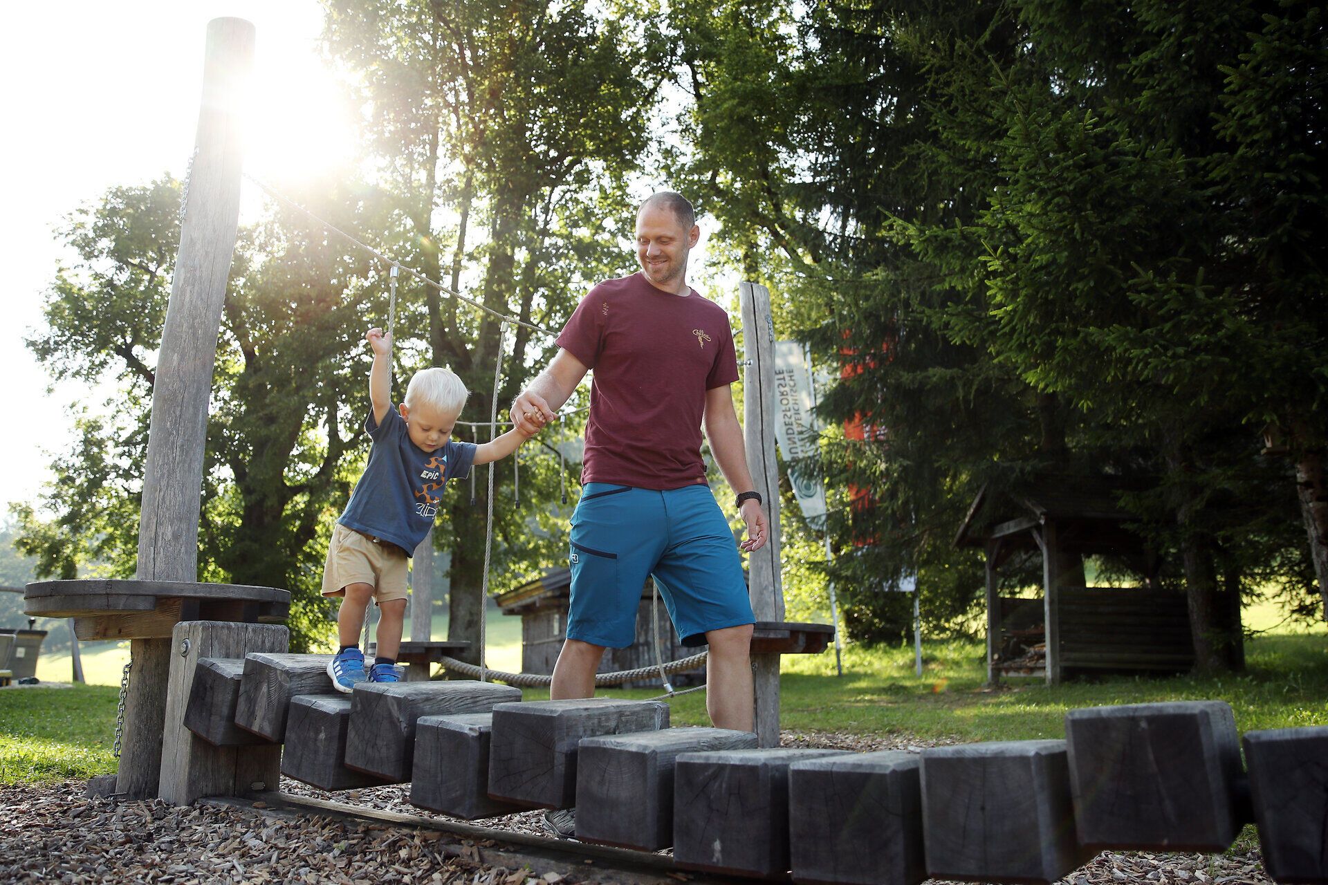 Ein Mann hält ein Kleinkind an der Hand während dieses über Holzblöcke am Spielplatz Promau balanciert. 