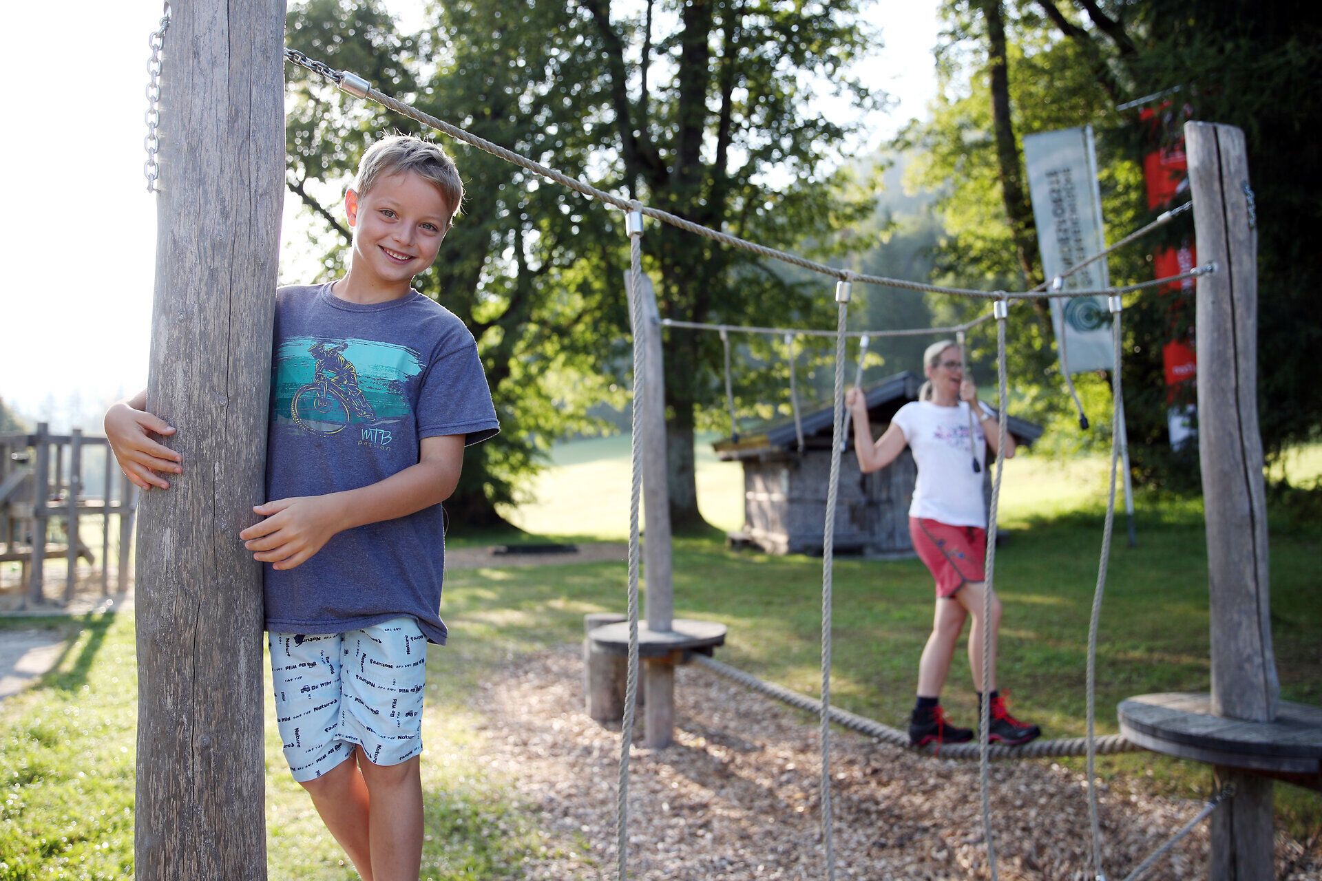 Der Junge im Vordergrund lächelt in die Kamera, während eine Frau im Hintergrund über ein Seil balanciert. 
