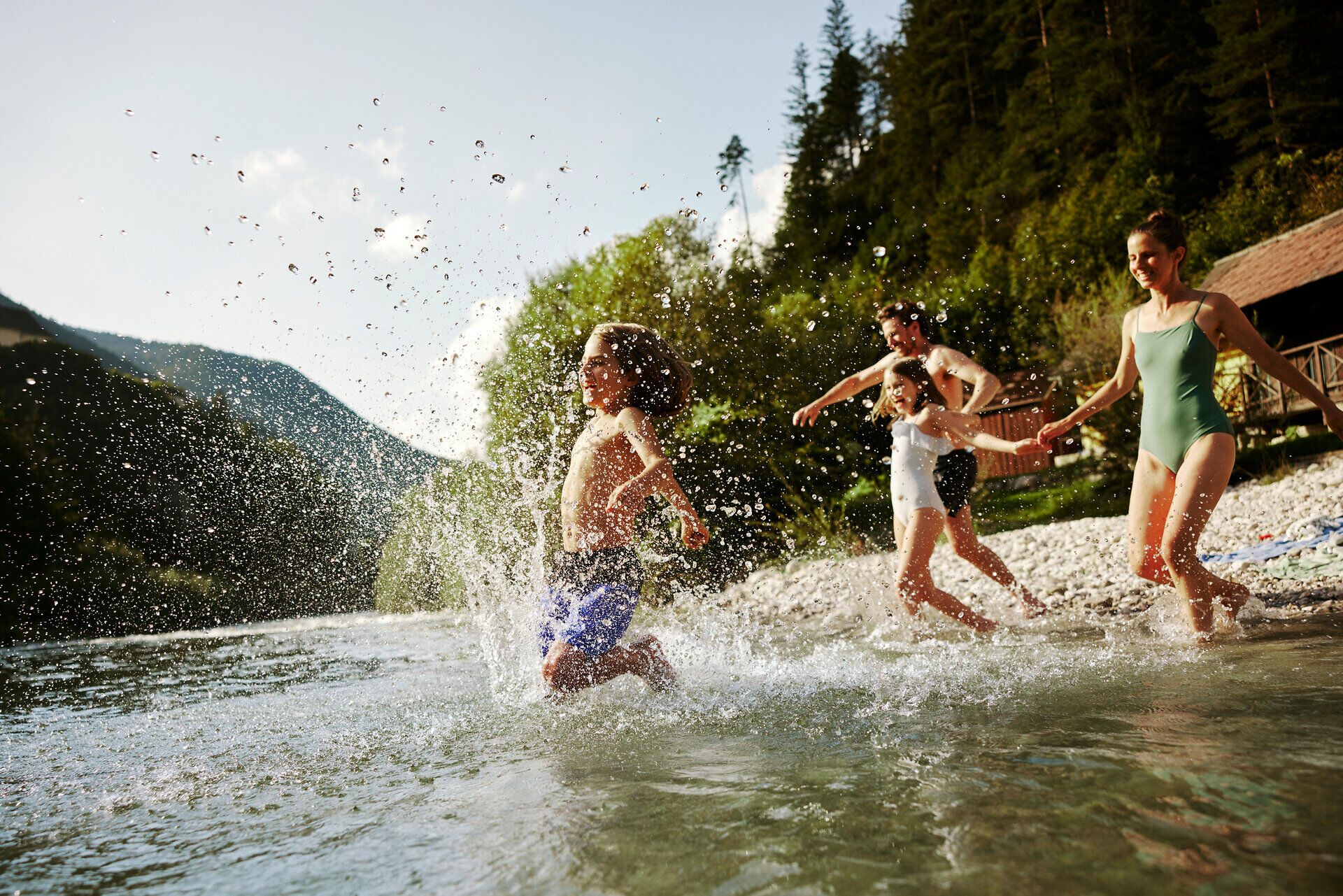 An einem sonnigen Tag genießen Kinder und Erwachsene das erfrischende Wasser eines klaren Flusses. Spritzende Wassertropfen und fröhliches Lachen schaffen eine unbeschwerte Atmosphäre, während die umliegenden Bäume Schatten spenden und die Schönheit der Natur unterstreichen.