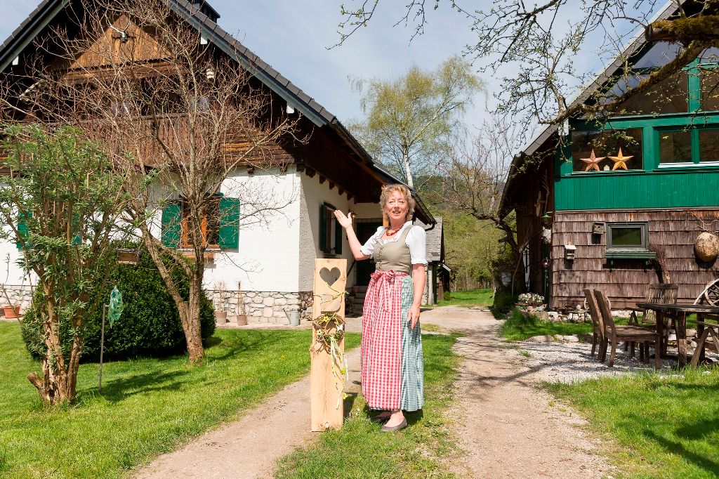 Eine Frau in traditioneller Kleidung steht vor zwei rustikalen Gebäuden auf einem grünen Hof.