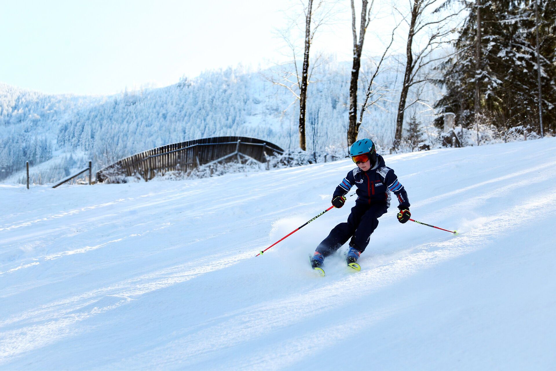 Ein fröhliches Kind gleitet mit Schwung die schneebedeckte Piste hinunter, umgeben von der malerischen Winterlandschaft. Die strahlende Sonne und der glitzernde Schnee schaffen eine einladende Atmosphäre für Familien, die die Freude am Skifahren entdecken möchten.