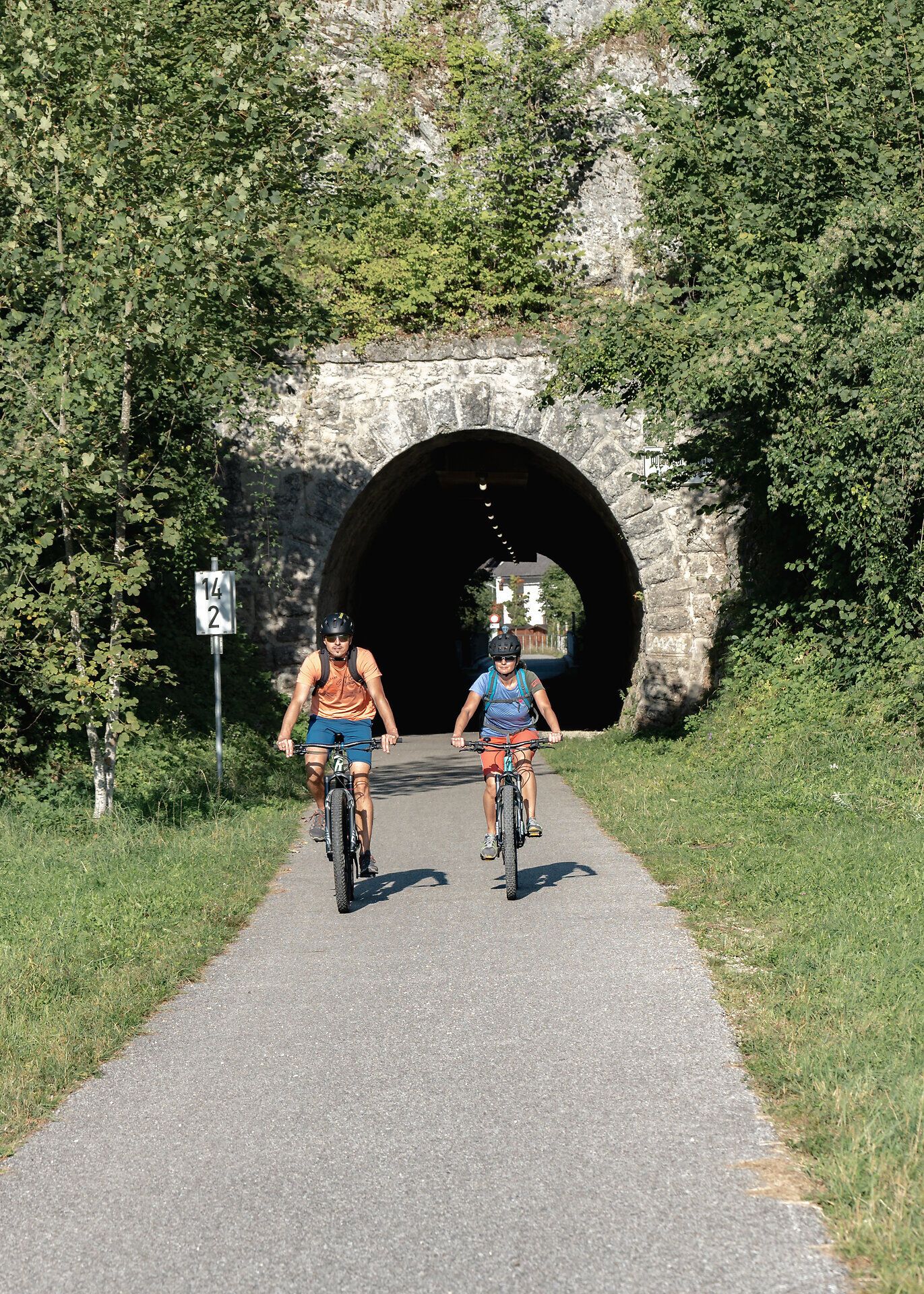Two cyclists enjoy the fresh air and picturesque surroundings of the Ybbstal Alps as they cycle through a shady tunnel. The lush vegetation and rolling hills create an inviting atmosphere for adventure seekers and nature lovers.