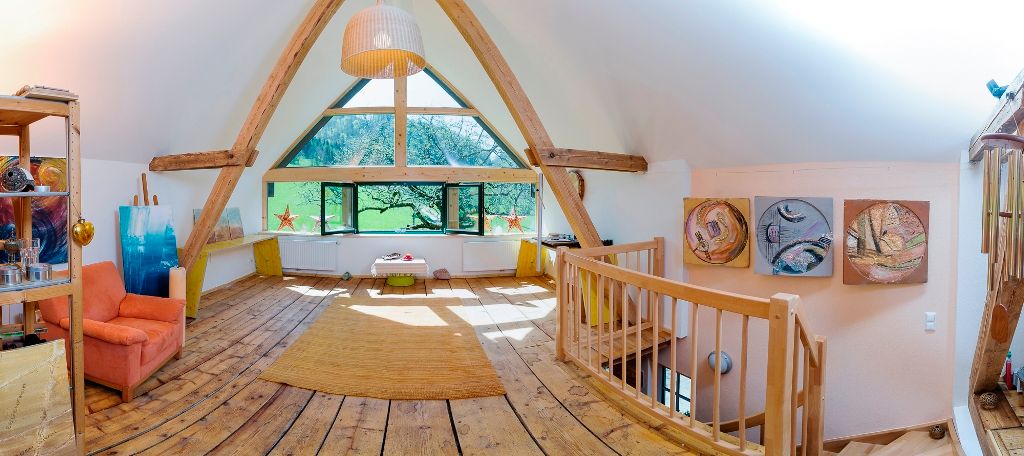 Innenansicht eines Dachgeschosses mit Holzboden, großen Fenstern und Kunstwerken an den Wänden.