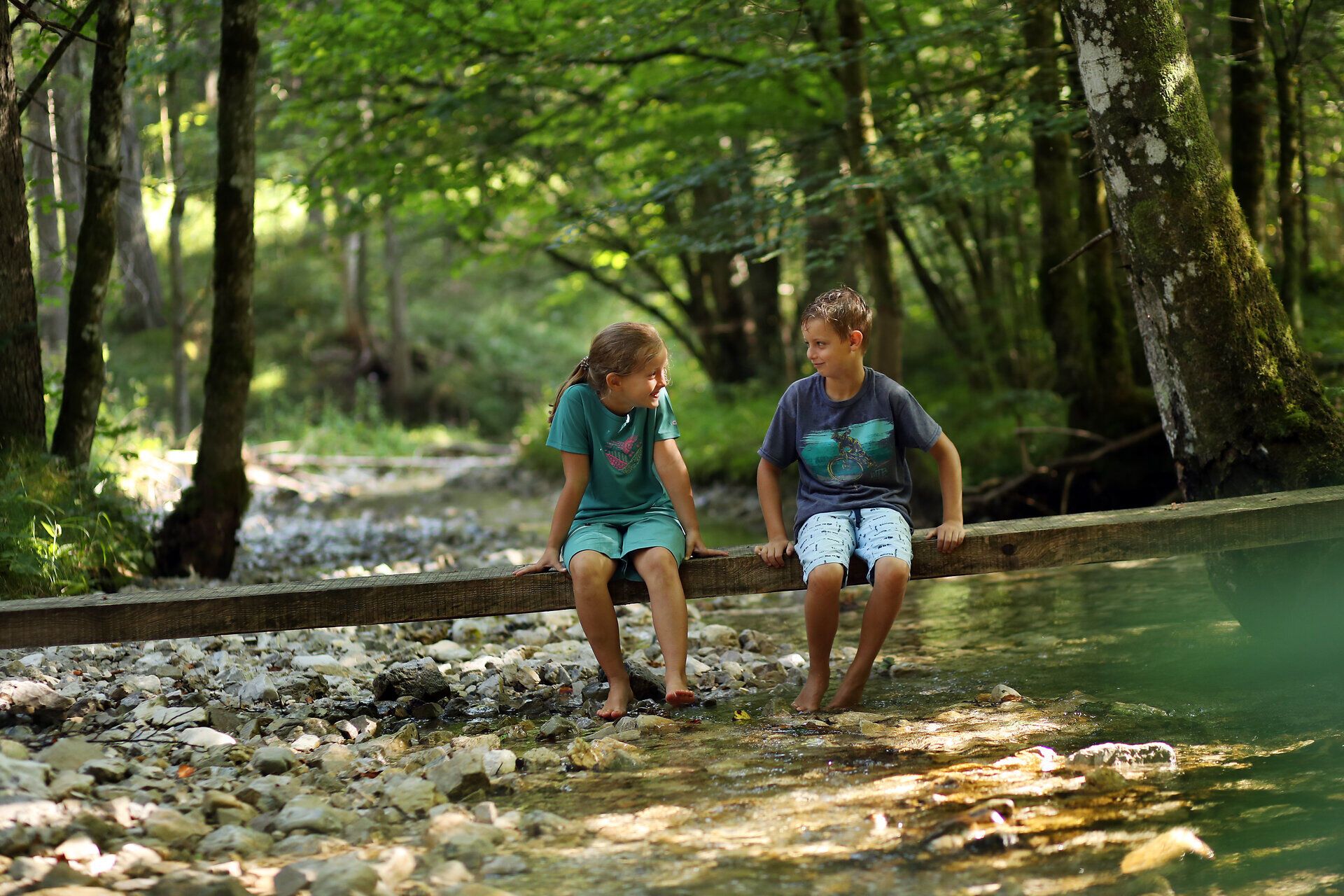 Kinder sitzen auf einem Balken über einem Bach im Naturpark NÖ Eisenwurzen.