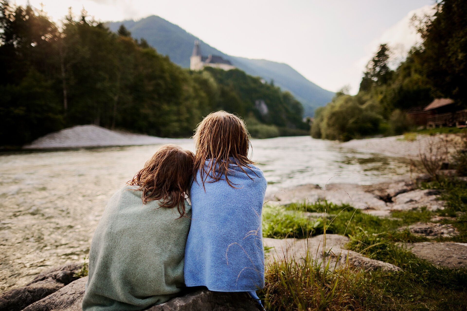 Zwei Kinder genießen einen unbeschwerten Moment am Fluss, eingehüllt in bunte Decken. Die sanften Wellen des Wassers und die umgebende Natur schaffen eine friedliche Atmosphäre, die zum Verweilen einlädt. Hier, wo die Berge auf das Wasser treffen, wird der Sommer in seiner schönsten Form erlebbar.