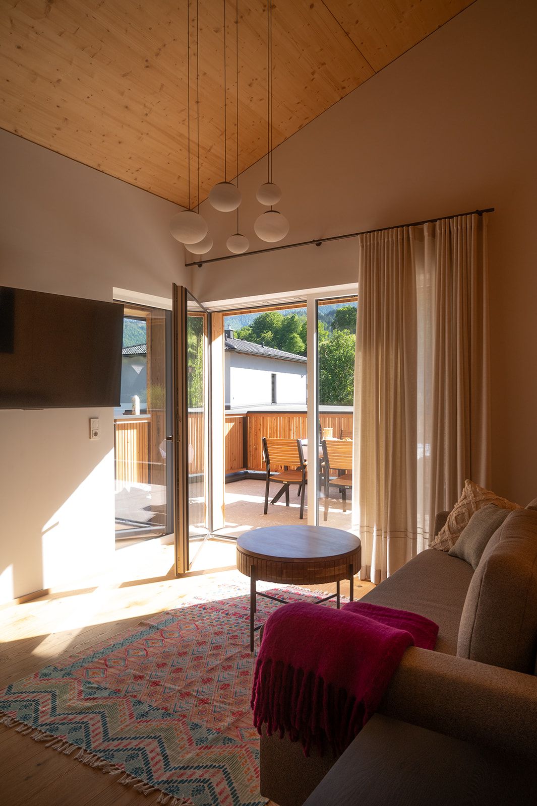 Interior view of a modern living room with wooden elements, a colorful carpet and access to a balcony with seating furniture.