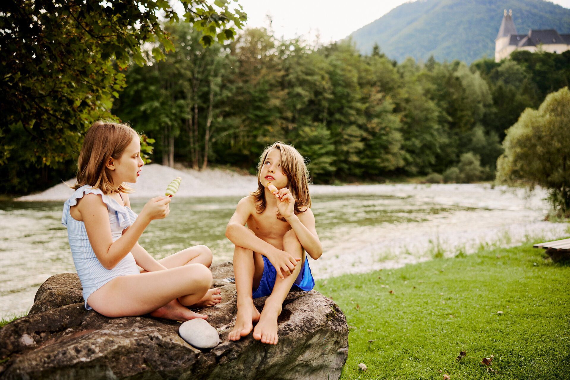 An einem sonnigen Tag genießen Kinder das erfrischende Wasser eines klaren Flusses, während sie auf einem großen Stein sitzen. Umgeben von üppigem Grün und sanften Hügeln, strahlt die Szene Freude und Unbeschwertheit aus, perfekt für einen Familienausflug.