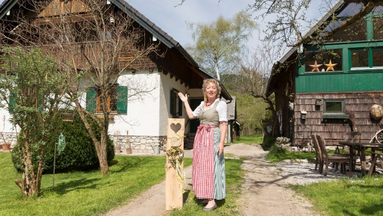 A woman in traditional dress stands in front of two rustic buildings in a green courtyard.