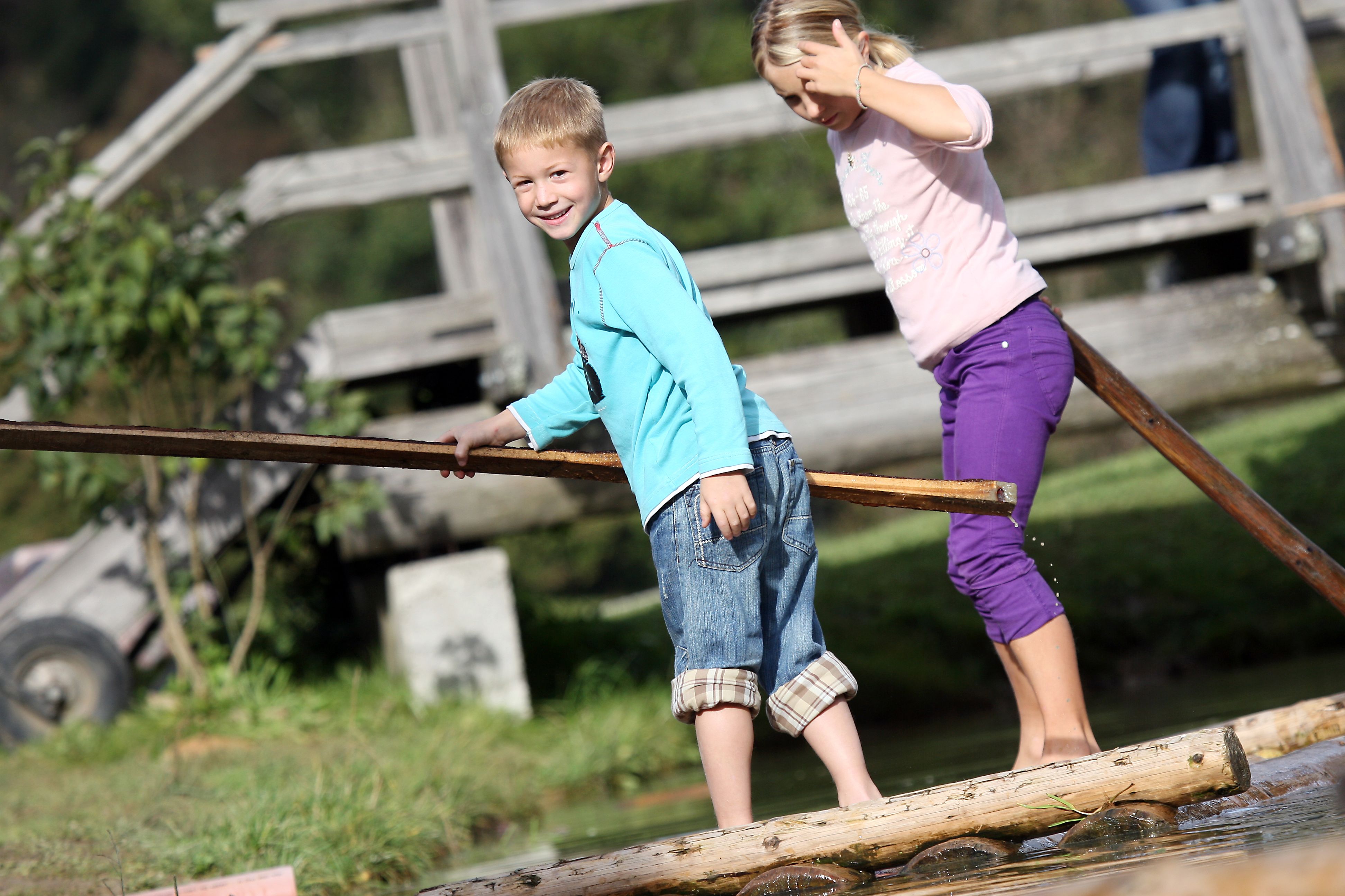 Zwei Kinder balancieren auf einem Holzfloß über Wasser, im Hintergrund eine Holzbrücke.