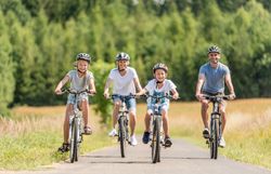 Eine fröhliche Familie radelt gemeinsam auf einem malerischen Weg, umgeben von üppigem Grün und sanften Hügeln. Die Sonne strahlt am blauen Himmel und schafft eine einladende Atmosphäre für unvergessliche Erlebnisse in der Natur.