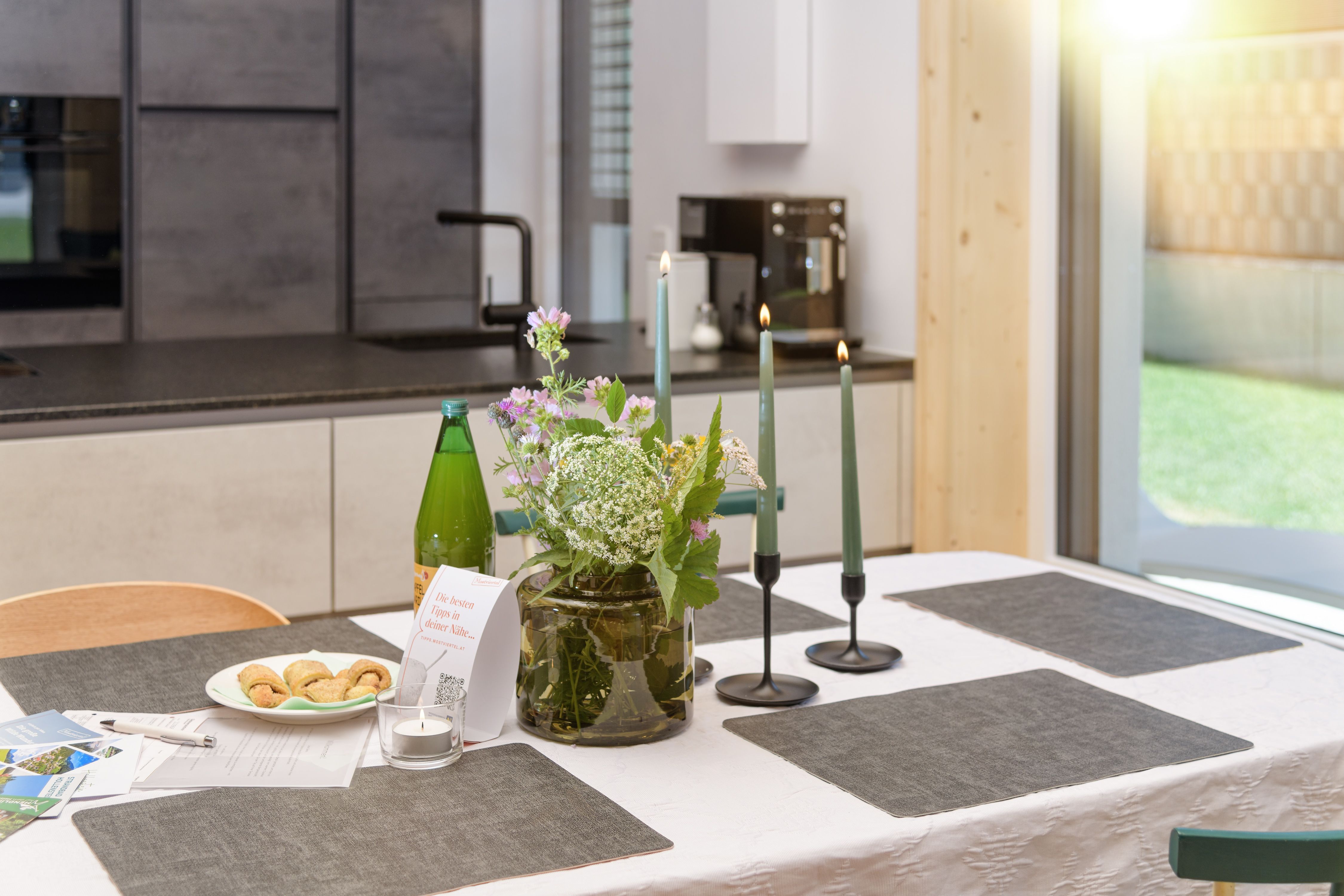 A set table in a modern kitchen with flowers, candles, and snacks.