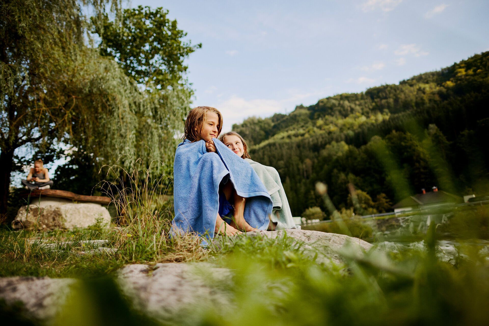 Zwei Kinder sitzen fröhlich am Ufer eines klaren Flusses, eingehüllt in bunte Handtücher. Die sanfte Brise und die umgebende Natur schaffen eine entspannte Atmosphäre, die zum Verweilen einlädt. Hier, wo die Schönheit des Mostviertels erlebbar wird, genießen sie den Sommer in vollen Zügen.