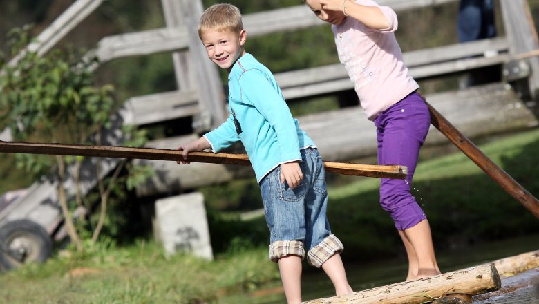 Zwei Kinder balancieren auf einem Holzfloß über Wasser, im Hintergrund eine Holzbrücke.