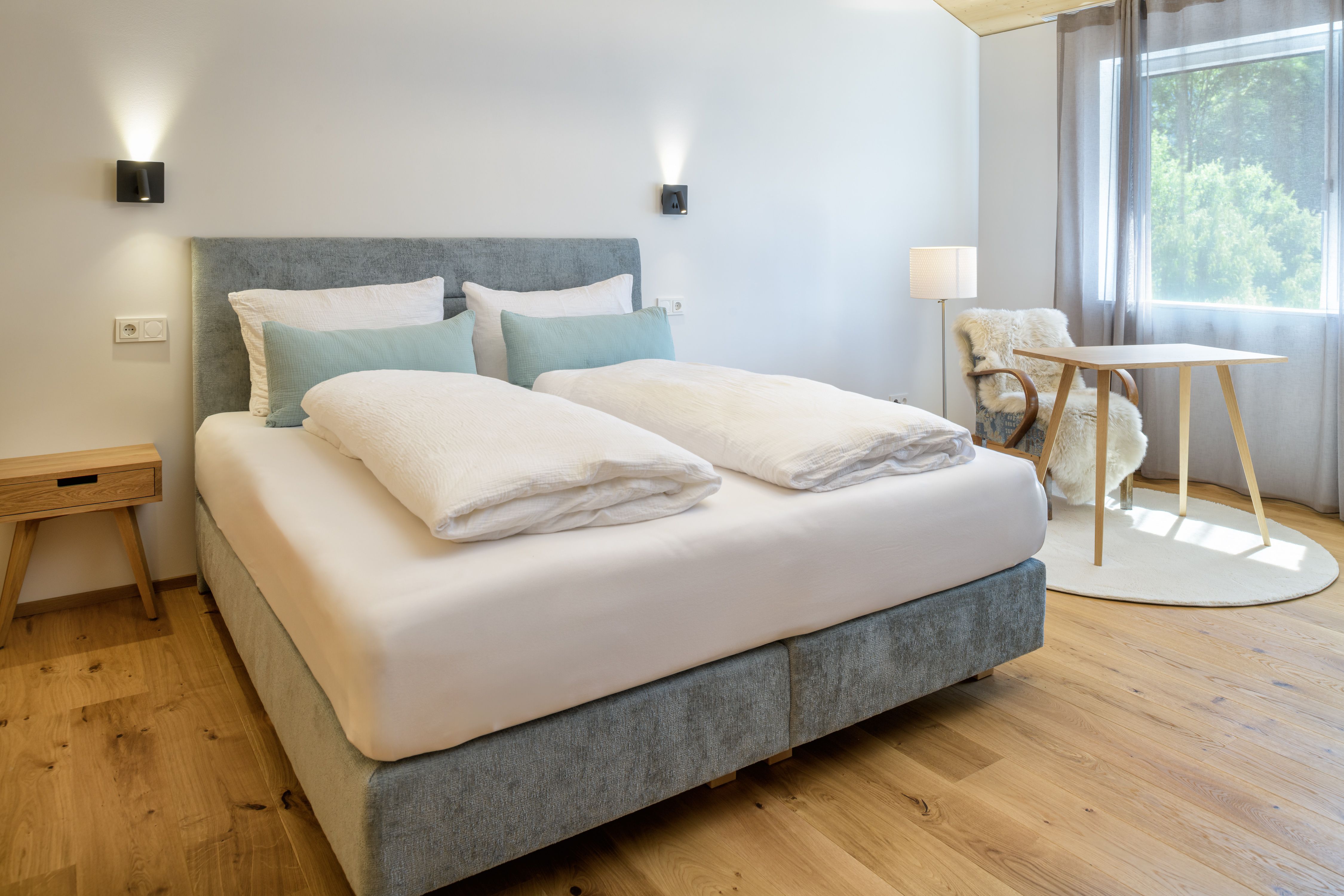 A modern bedroom with a large bed, hardwood floors, a small table, and a chair with a fur throw. Natural light streams in through a window.