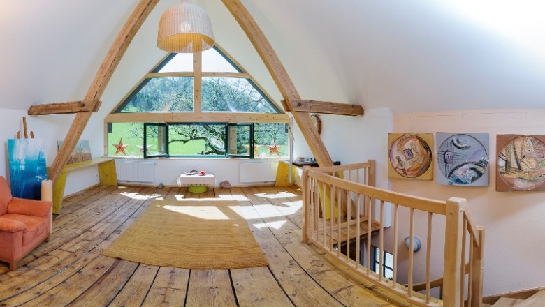 Innenansicht eines Dachgeschosses mit Holzboden, großen Fenstern und Kunstwerken an den Wänden.