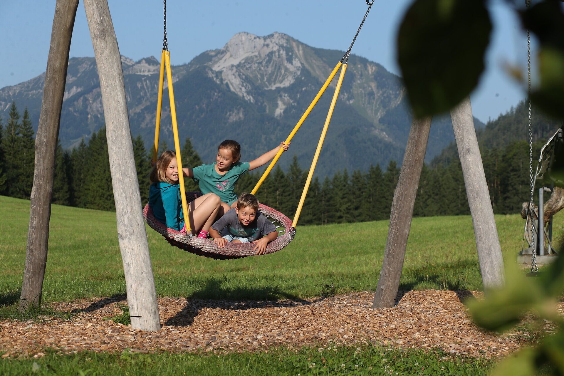 Drei Kinder sitzen bei Sonnenschein auf einer Korbschaukel, im Hintergrund sieht man die Voralpe. 