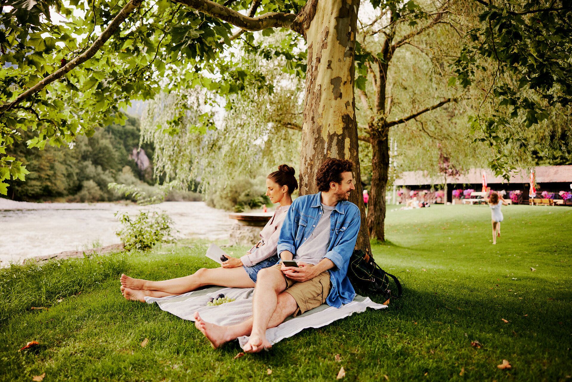 Unter dem schattenspendenden Baum genießen eine Familie und ihre Kinder die entspannte Atmosphäre am Fluss. Das sanfte Plätschern des Wassers und das Lachen der Kinder schaffen eine harmonische Stimmung, die zum Verweilen einlädt.