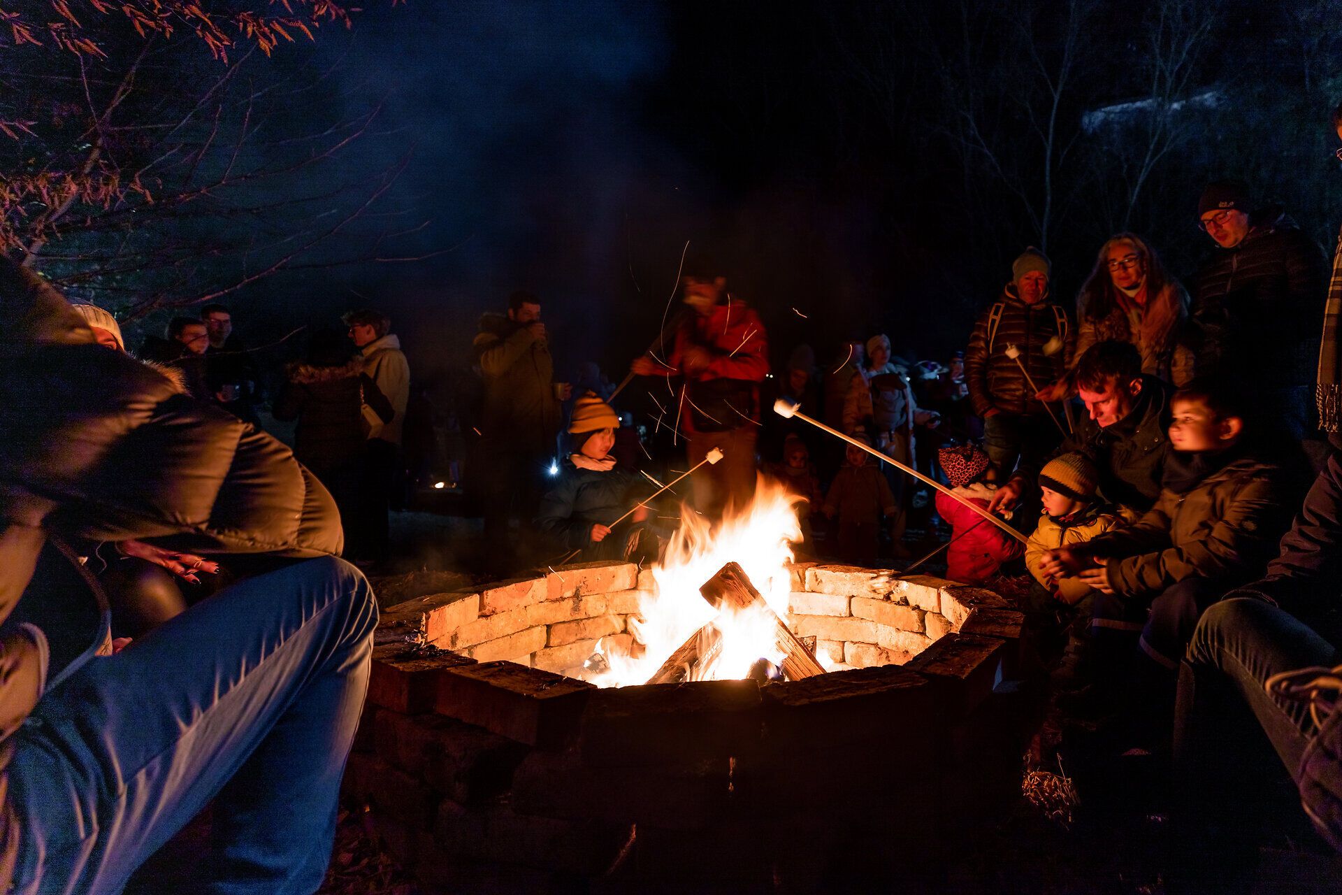 Menschen sitzen um ein Lagerfeuer auf einem Weihnachtsmarkt bei Nacht.
