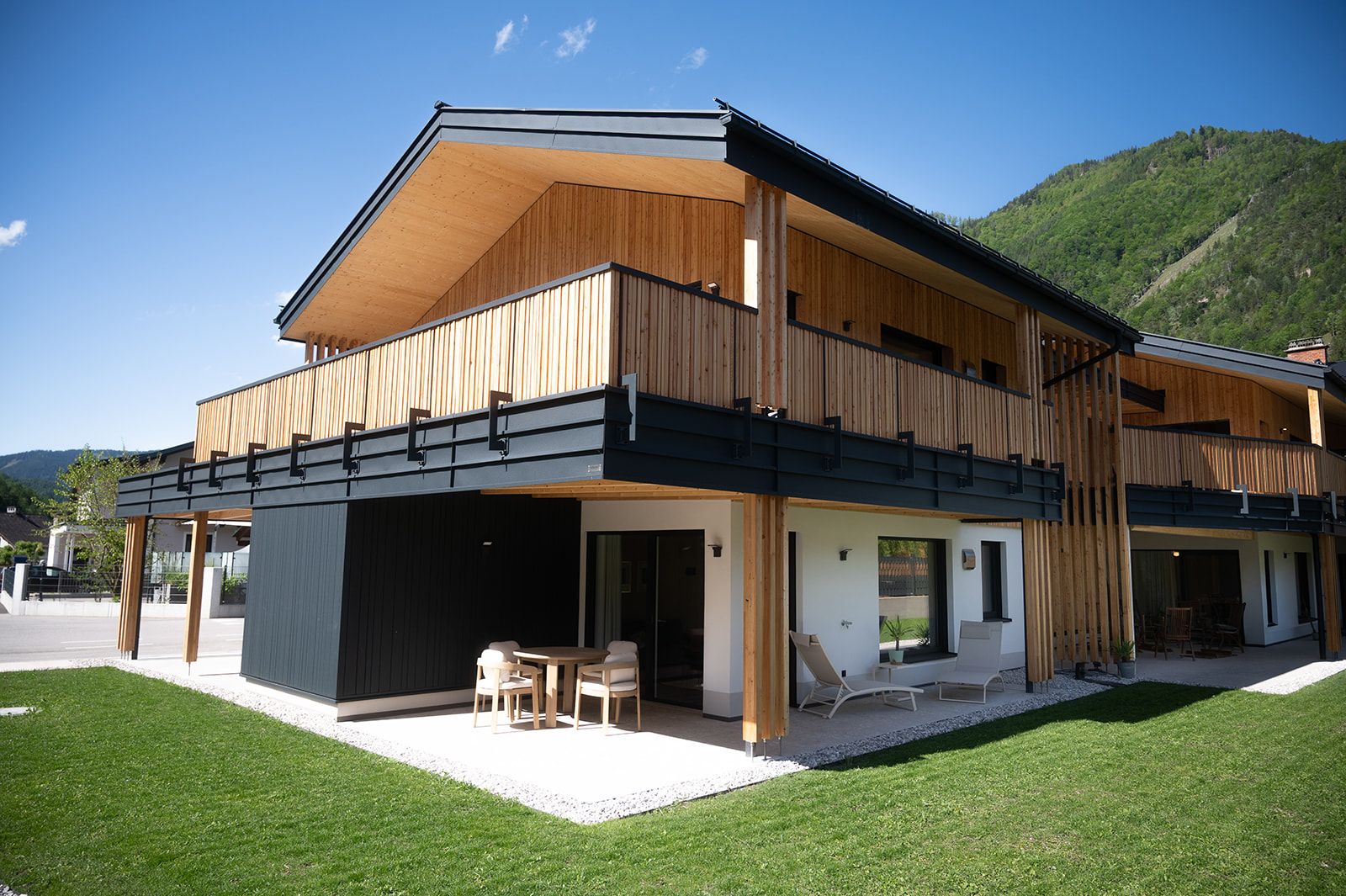 Modern wooden architecture of a two-storey building with balcony and terrace, surrounded by green landscape.