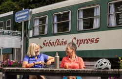 Zwei fröhliche Wandererinnen genießen eine wohlverdiente Pause auf einer Bank, während sie die frische Bergluft und die malerische Aussicht auf die umliegenden Hügel und Wälder aufnehmen. Die strahlende Sonne und die bunten Blumen um sie herum schaffen eine einladende Atmosphäre, die zum Verweilen einlädt.