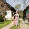 Eine Frau in traditioneller Kleidung steht vor zwei rustikalen Gebäuden auf einem grünen Hof.